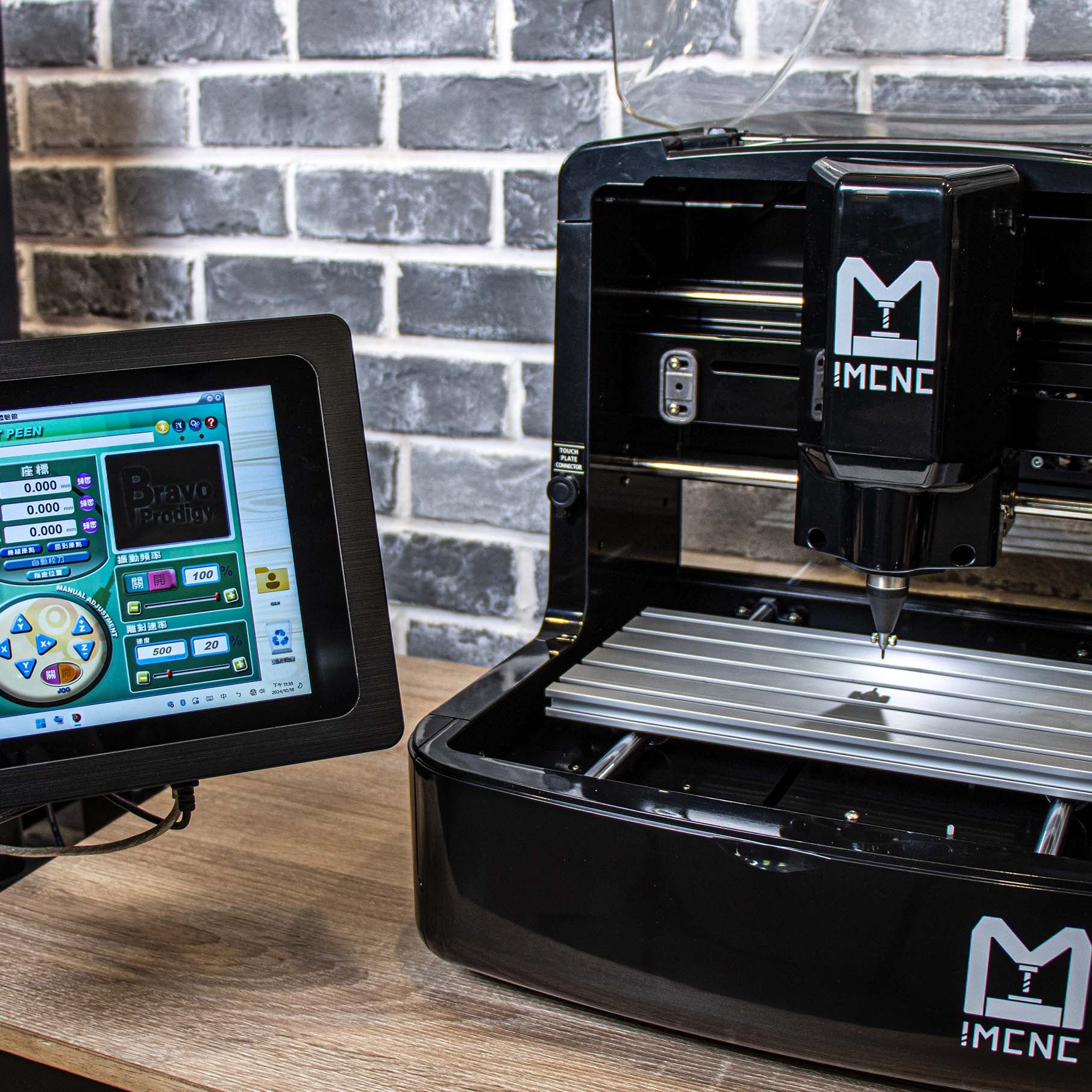 Mini desktop CNC engraving machine in Dot peen marking module by IMCNC, with touchscreen controller displaying CNC software interface, placed on a wooden table against a brick wall background.
