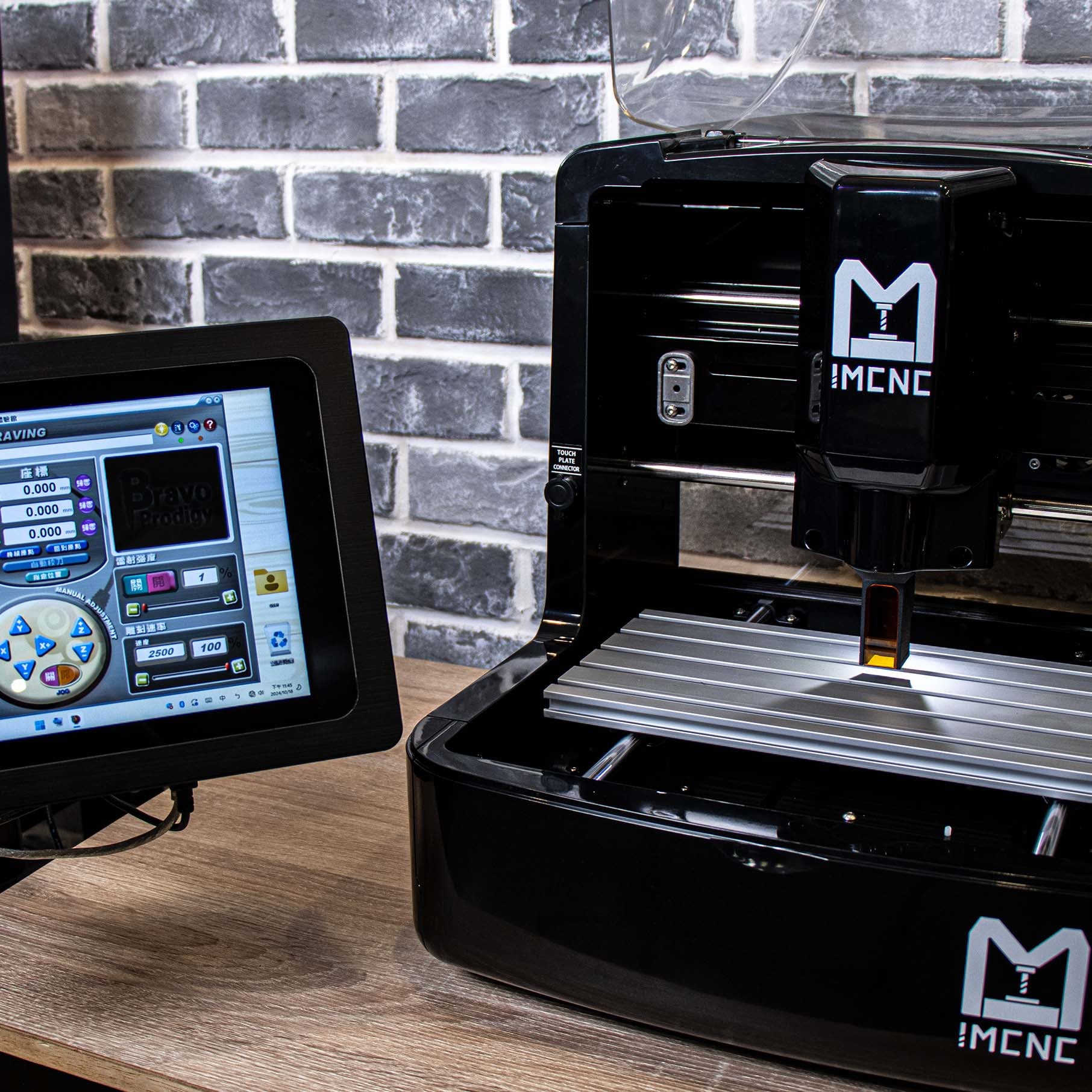 IMCNC mini desktop CNC engraving machine with laser module, connected with touchscreen controller, shown during operation on a metal work surface, placed on a wooden table with brick wall background.