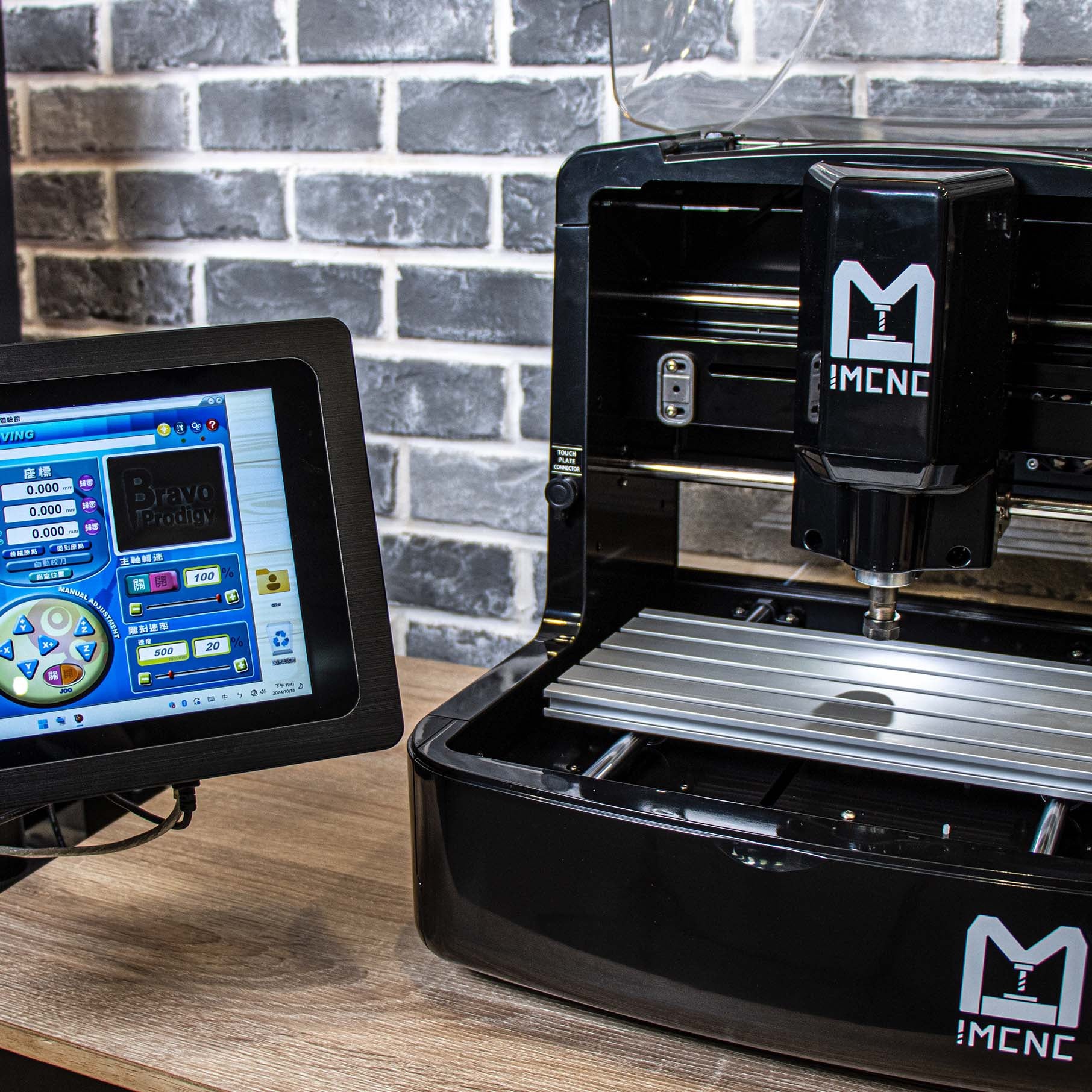 Compact desktop CNC engraving machine with IMCNC logo, shown alongside a touchscreen control panel displaying CNC software interface, set against a brick wall background.