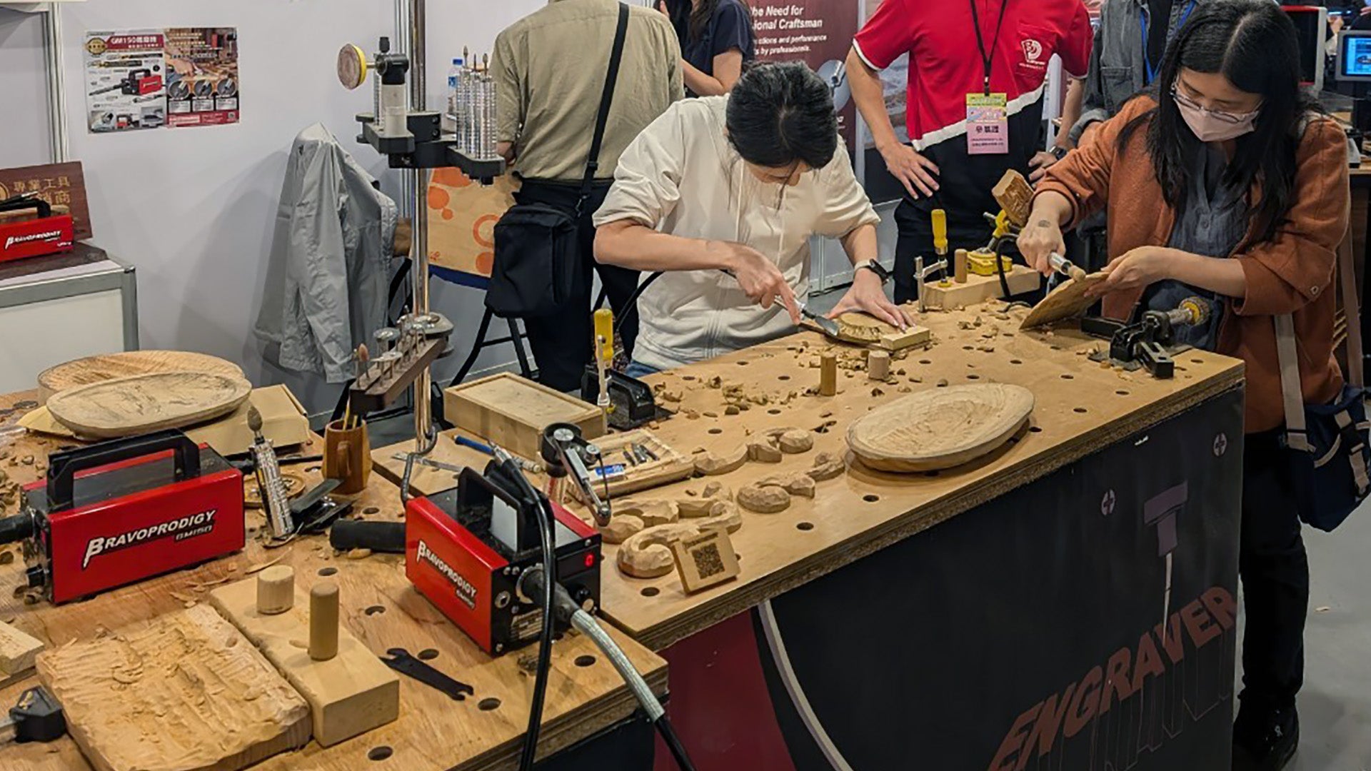 People at a public booth carving wood on a large workbench filled with GM150 Flexible Shaft Grinder, rotary engravers, and wooden art pieces.