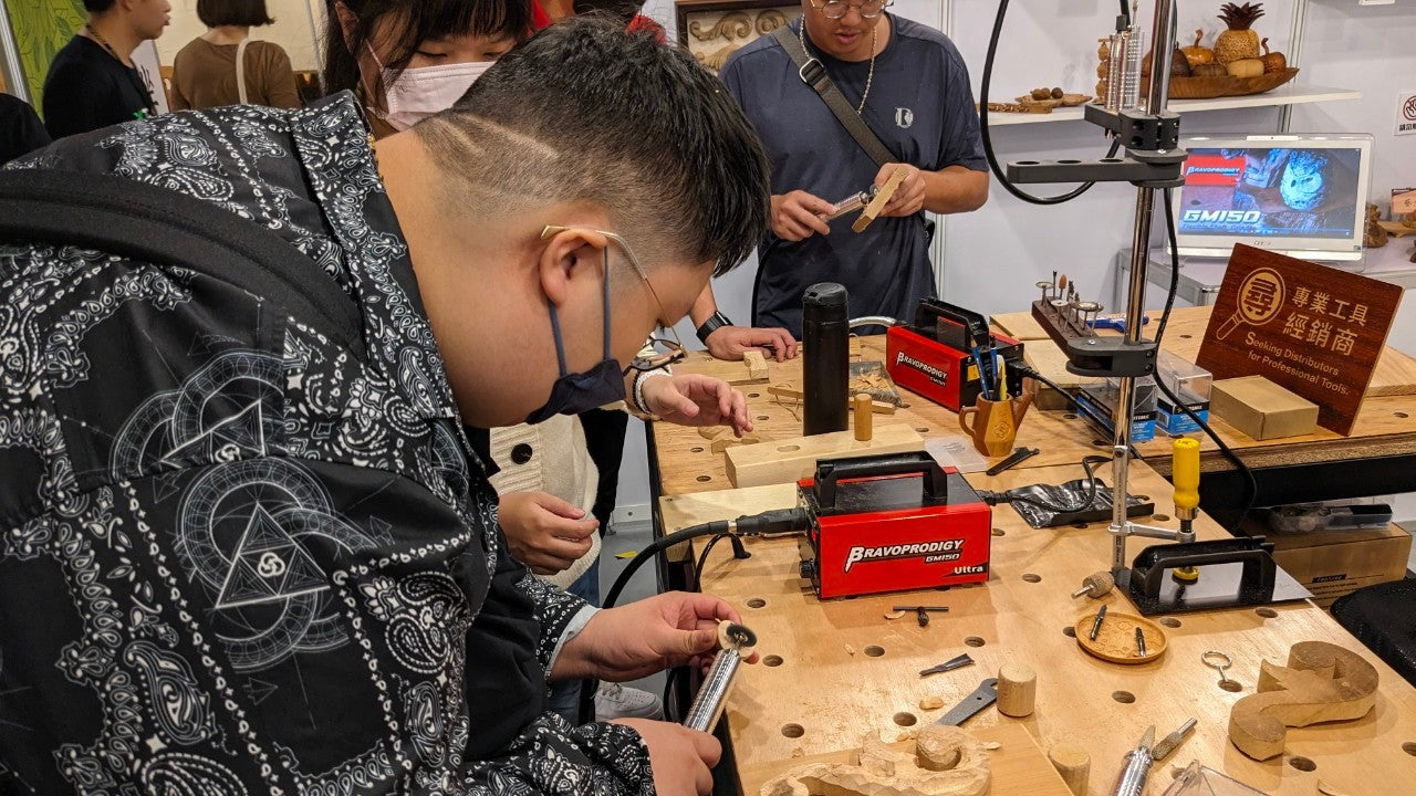 A group of people gathered around a workbench at a CNC demonstration booth, with one participant focusing on a handheld rotary tool connected to a red BRAVOPRODIGY Flex Shaft Grinder.