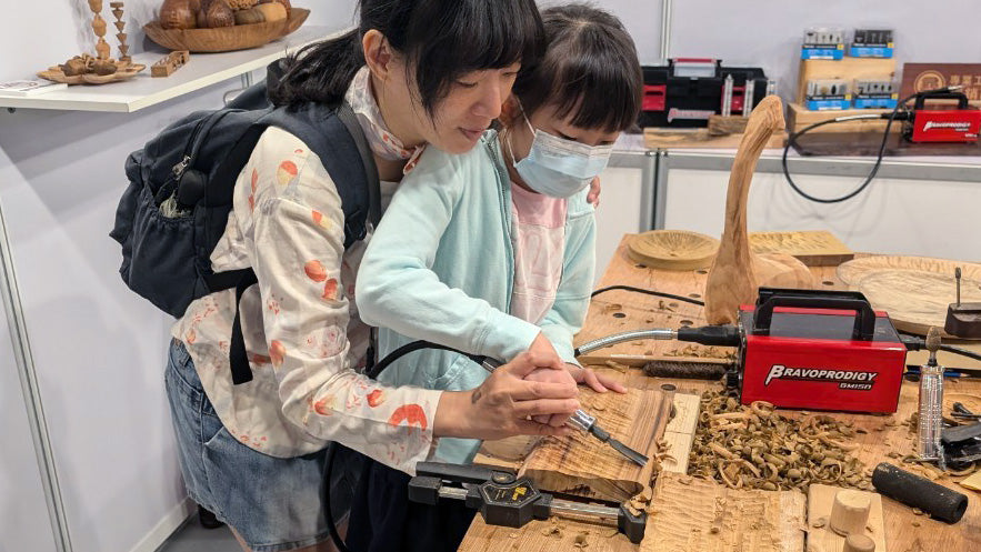 A woman guiding a child in using a rotary carving tool on a wooden board during a CNC woodcarving demonstration, with a BRAVOPRODIGY Flex Shaft Grinder visible in the background.