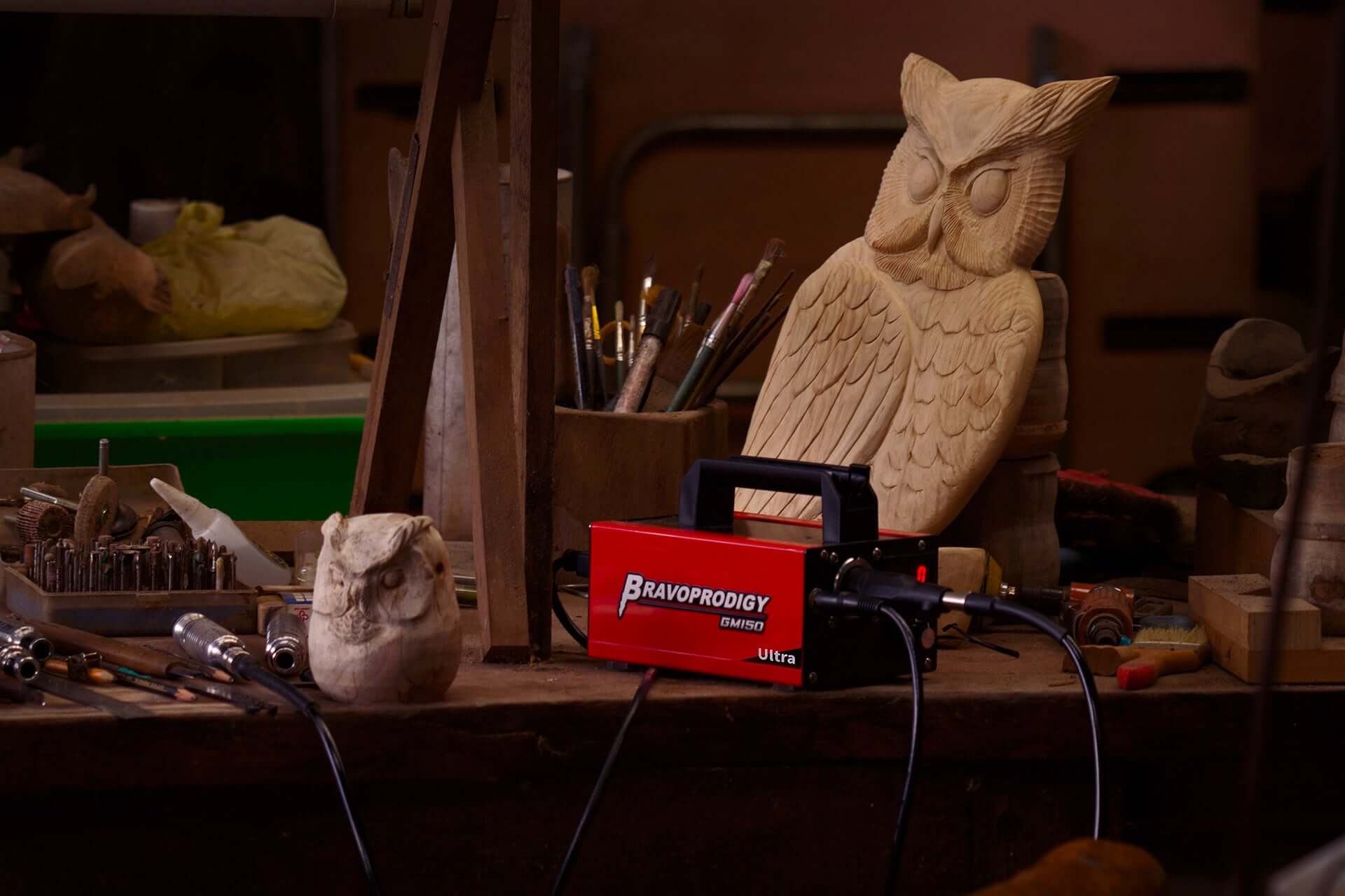 A wooden owl sculpture in progress, surrounded by carving tools and a red BRAVOPRODIGY CNC Flex Shaft Grinder on a cluttered workbench.